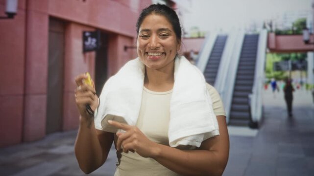 Woman holding stopwatch, finger pointing to timer with towel draped over shoulders in building entrance, smiling and checking time; fitness motivation confidence.