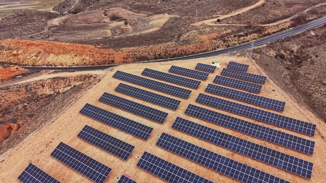 The setting shows many solar panels arranged in rows on a hillside. The panels face the sun and are surrounded by natural land. A road runs along the edge of the area.