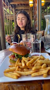 Vertical format of a young girl sitting on the patio of a rustic cabin style restaurant, eating something spicy and having some funny facial reactions. A stacked burger and fries in view.