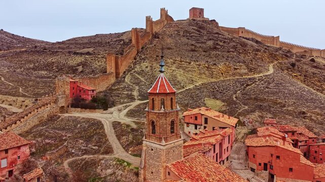 A village with red rooftops sits at the base of a large castle on a hill. The castle has ancient walls that stretch across the landscape. The sky is cloudy, and the area is rugged.