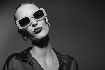 Stylish white woman with fair skin wears large sunglasses and hoop earrings, posing confidently in a dark satin shirt in black and white portrait.