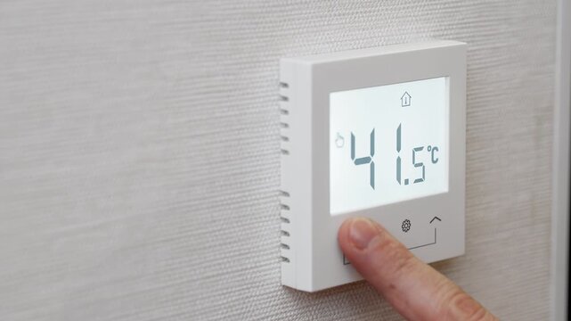 Close up of hand adjusting digital thermostat showing high temperature on smart home wall controller. Concept of heating system climate control and home energy management.