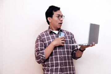 Man with glasses looks surprised while holding phone and laptop. He wears a plaid shirt and stands...