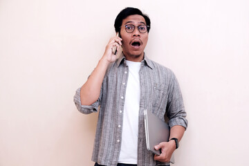 Man with glasses looks surprised while holding phone and laptop. He wears a plaid shirt and stands against a plain background. His wide eyes and open mouth show shock or excitement © Nurma