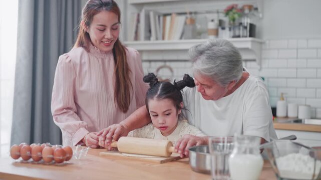 Grandmother teaches her granddaughter to roll dough while the smiling mother watches, creating a heartwarming three-generation baking moment in a kitchen.