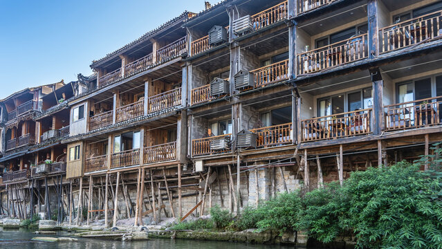 An old wooden building on stilts on the riverbank. Balconies with railings. Green vegetation on the shore. China. Fenghuang Ancient Town.  Phoenix. Tuojiang River 
 