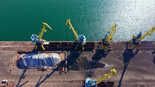 Cranes are positioned along the dock, moving cargo items at a shipping terminal. The bright blue water reflects the work happening above as trucks wait nearby.