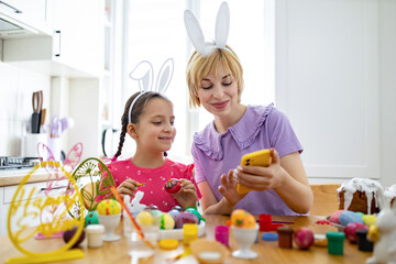 Obraz premium A mother and her daughter wearing bunny ears look at a smartphone while decorating Easter eggs together at a table