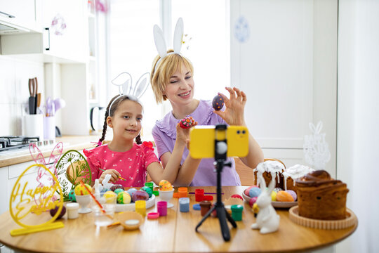 A mother and daughter wearing bunny ears are decorating Easter eggs and recording a video on a smartphone