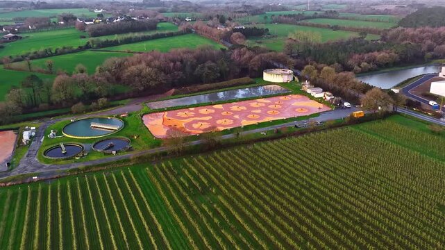Drone aerial view of colorful oxidation ponds and settling tanks with orange floral patterns at wastewater treatment facility in Jelgava Latvia surrounded by farmland and forest