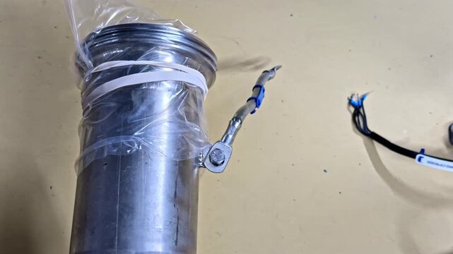 Aluminum stainless steel cylindrical sensor housing wrapped in plastic film with attached temperature probe cable blue zip ties and crimped terminals in industrial assembly setup