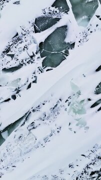 Top-down drone view of cracked sea ice covered in windblown snow streaks, with turquoise water patches forming an abstract winter texture background. Vertical.