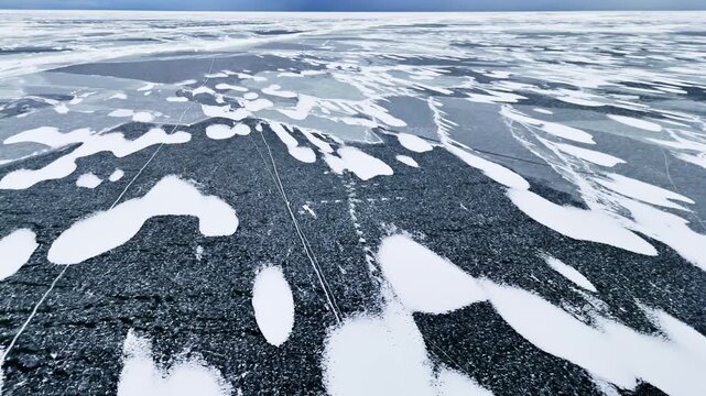 Aerial glide over patterned frozen sea ice with scattered snow patches and long crack lines, creating abstract winter textures across a vast cold landscape.