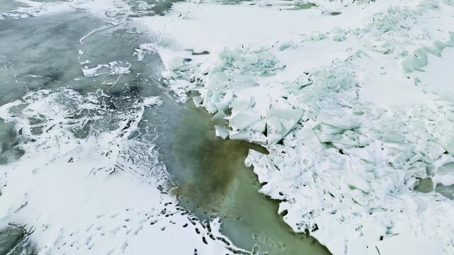 Aerial view gliding over a frozen sea pressure ridge with pale green meltwater channel, broken ice blocks, snow patches, and dangerous unstable winter ice.