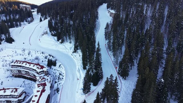 Aerial view shows a winding skiing trail surrounded by tall trees and buildings covered in snow. The scene captures winter activities in a remote area during daytime.