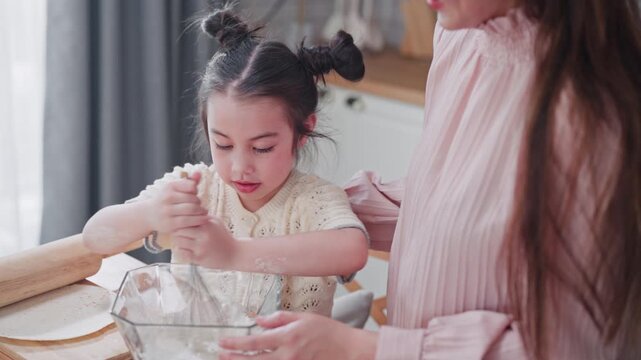 Focused young Asian girl earnestly whisks batter in a glass bowl while her mother assists nearby, with a rolling pin and flour visible on the kitchen table.