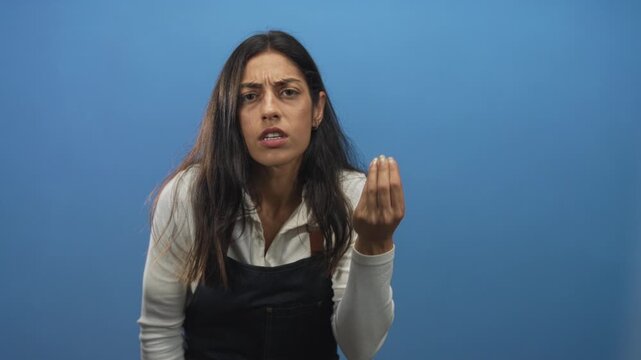 Woman with pinched fingers gesture and tense face in blue studio leaning forward wearing dark apron; frustration insistence urgency.