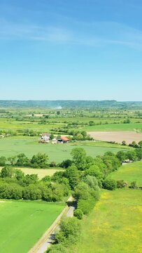 Aerial view of the lush green fields, farmland, and scattered trees in the Normandy countryside on a clear summer day. Rural houses and agricultural plots are visible under a bright blue sky.