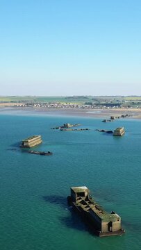 Aerial view of concrete remnants of an artificial harbor in the turquoise waters off Gold Beach near Asnelles, Normandy, with a coastal village and fields in the background under a clear blue sky.