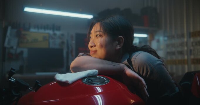 Tired But Happy Female Biker Resting Her Head on Motorcycle Tank After Repair and Polishing. Atmospheric Moment in Garage Workshop. Concept of Peace and Accomplishment in Her Private Sanctuary.