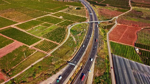 The view shows a busy road curving through fields of crops and vineyards with different colors and textures during daytime. Vehicles are moving along the roadway.