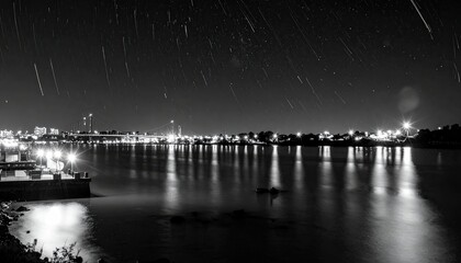 Fototapeta premium City lights shimmer over water with streaking stars above in black and white photo