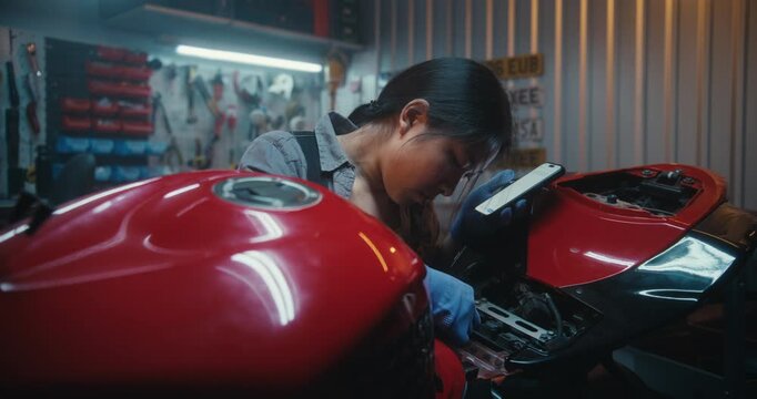 Young Woman in Workshop Consults Artificial Intelligence Voice Assistant During Complex Motorcycle Repair. Girl Studies Exposed Components, Reconnects Cables, Battery Step By Step With Guided Support.