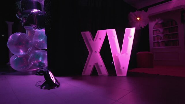 Reveal shot of illuminated XV letters decoration with balloons and pink lighting arranged for quincea&ntilde;era birthday celebration event.