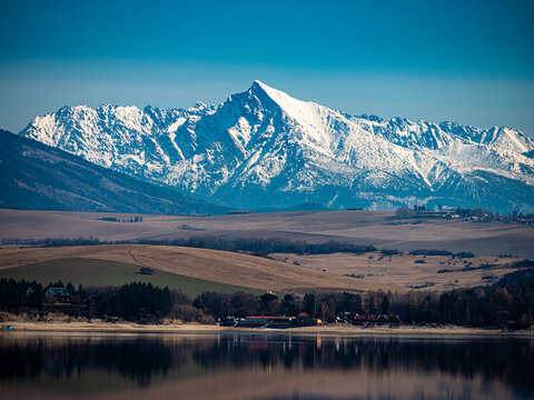 Kriv&aacute;ň peak in High Tatras reflected in calm water of Liptovsk&aacute; Mara reservoir with rolling Liptov landscape in foreground. Scenic mountain panorama in northern Slovakia.