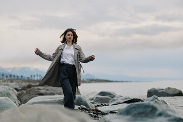 Young woman wearing long coat walks on rocky shore near calm sea under cloudy sky. Concept of...