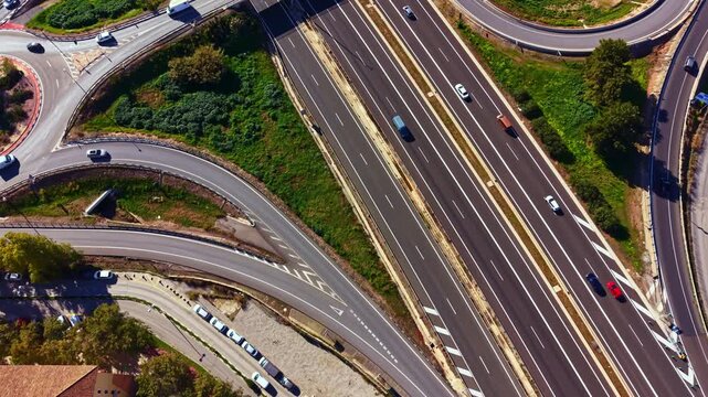 Cars drive on various lanes of a highway interchange in a city. Some vehicles move toward exits while others continue on the main road. Green areas are visible along the sides.