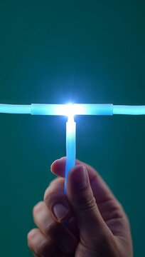 Close-up of a hand holding a blue glow stick, connecting it to two other glow sticks, creating a bright, glowing T-shape against a dark background.
