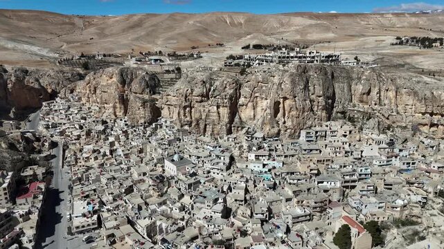 A wide aerial drone panorama of Ma'loula, Syria, showcasing the ancient village built into the massive rocky cliffs of the Qalamoun Mountains