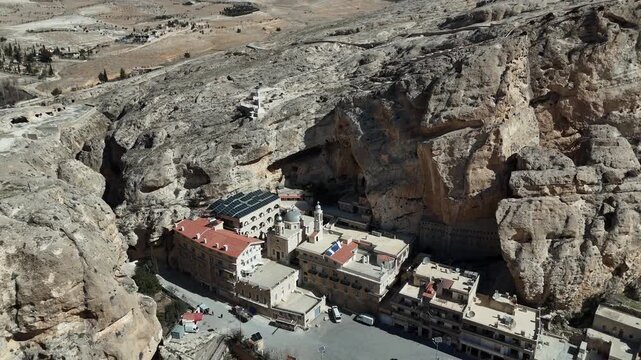 Drone shot flying close to the historic stone houses of Ma'loula, Syria, nestled directly beneath the towering, sheer rocky cliffs of the Qalamoun Mountains