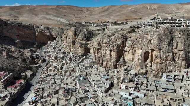 Wide aerial panorama of Ma'loula, Syria, captured from the surrounding arid slopes, showing the ancient village spread along the base of massive desert cliffs