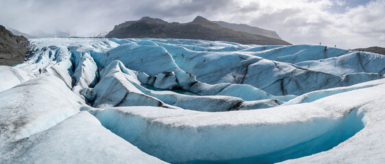 Panoramic view of a massive blue glacier with deep crevasses and icy ridges under a cloudy sky, featuring tiny figures of hikers for scale in a majestic arctic mountain landscape in Iceland. © Nazran