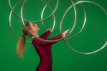 Side view of a young circus gymnast in a red leotard standing on a green background and holding the hoop on her forehead, and spinning it on her arms. © kinomaster