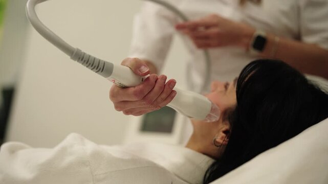 Tracking shot of relaxed middle-aged female client receiving microneedle radiofrequency face lifting procedure from dermatologist for skin tightening and rejuvenation in professional aesthetic clinic.