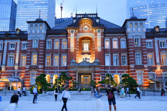 A historic red brick public building illuminated at dusk, contrasting with modern glass skyscrapers in the background while commuters walk in the plaza