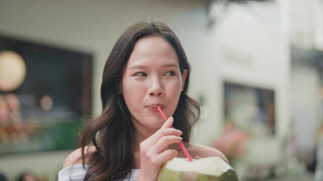 Close-up of a relaxed young Asian woman drinking fresh coconut through a straw outside, capturing a natural and refreshing summer moment.