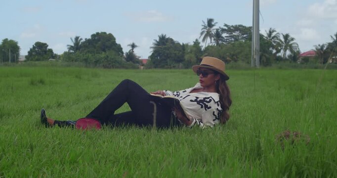 A girl in a tailored maroon pantsuit and a matching felt hat reads a book on a park blanket with a panoramic mountain view.