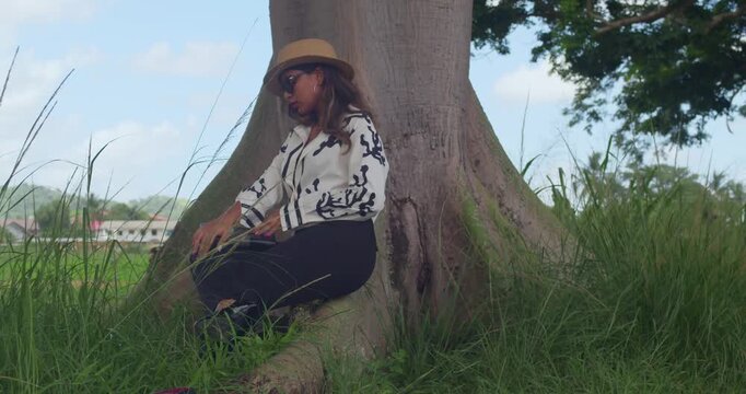 Professional attire meets nature as a girl in slim-fit slacks and a felt hat reads a hardcover book in a sunny valley park.