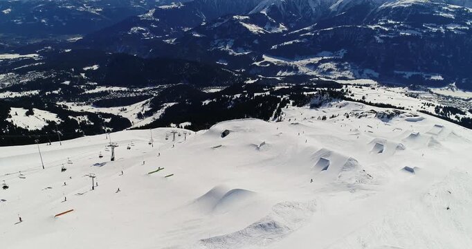 High aerial drone shot overlooking the LAAX snowpark with jumps, rails, and groomed lines visible across the slope. Wide alpine panorama and cinematic winter sports landscape.