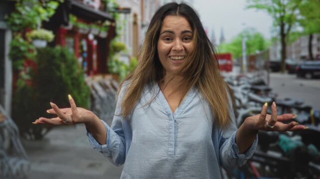 Woman shrugs with open hands and windblown hair on a european street lined with scooters and storefront planters; playful surprise.