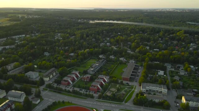 Drone aerial rising above a green suburban neighborhood with houses, roads and distant smoke drifting across the forest on a sunny summer morning.