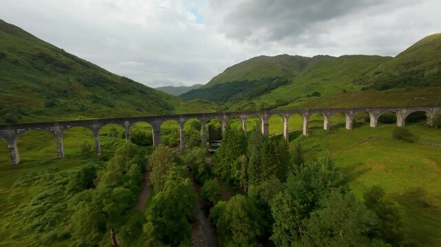 FPV drone glides over the empty, historic Glenfinnan Viaduct, showcasing the vast arches and deep blue waters of Loch Shiel. This bridge is the famous route of a steam train featured in wizarding lore