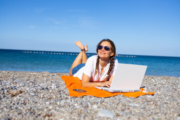 Young woman working remotely on laptop while lying on beach towel by the sea