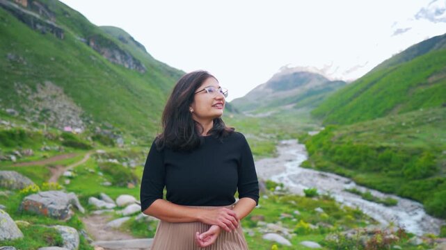 Portrait of Indian woman looking at mountain valley and flowing river in Sural Bhatori, Pangi Valley, India. Happy tourist exploring hidden gems. Summer mountains with snow peaks and dark clouds in sk