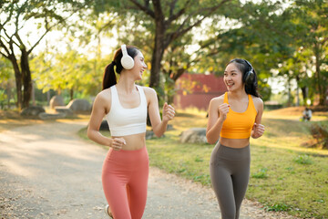 Fototapeta premium Two active women jogging together in a green park while listening to music with headphones, enjoying outdoor fitness, healthy lifestyle, friendship and energetic morning workout.
