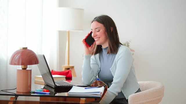 A young woman is deeply engaged in a focused phone conversation while simultaneously working on her laptop at a tidy desk. She appears attentive and productive as she jots down notes.